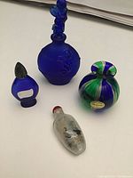 Four perfume bottles on white surface including cobalt blue cherub bottle, Murano bottle, reverse painted snuff bottle, and Evening in Paris bottle