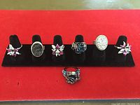 Seven sterling silver rings displayed on black velvet stand with red background. Includes floral and oval druzy style stones, all faux.