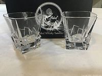 Two clear square-bottom whisky tumblers displayed in front of branded box
