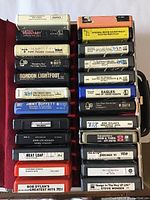 Top view of 24 vintage 8-track tapes stacked in groups of six, showing labels with various artists and titles including Meat Loaf and Bob Dylan