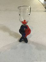 Front view of the Murano clown shot glass showing clear glass top with painted eyes and bright red lips, and figurine base in blue and red outfit standing with crossed legs.