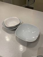 Four white ceramic bowls in varying sizes, square shaped with rounded edges, placed on a kitchen counter, showing general signs of wear and use.
