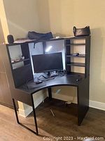 Front view of black IKEA corner study desk with visible signs of wear, displaying shelving units and desk surface with a few electronic items placed.