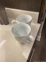 Two white ceramic mugs on a kitchen countertop, showing the open tops and handles.