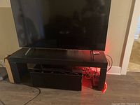 Photo of a black TV stand with a glass top, metal legs, holding a flat screen TV, with visible damage to the surfaces and edges.