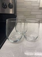 Three clear stemless wine glasses on a white countertop with a kitchen stove and tile backsplash in the background.