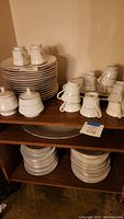 Photo of stacked dinner plates, cups, saucers, and other pieces on wood shelving showing the quantity and arrangement.