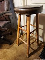 Photo of a wooden bar stool showing its round brown leather padded seat and natural wood legs with cross supports.