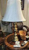 Brass table lamp with white bell-shaped fabric lampshade set on a round wooden table, with other items nearby.