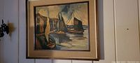 Framed painting showing harbor scene with boats and buildings, viewed straight on against light wall.