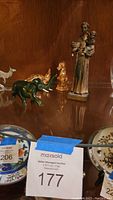 Jade elephant, pewter kangaroo, gold ducklings, monk holding child figurine shown inside a cabinet shelf with part of decorative porcelain plates visible below.