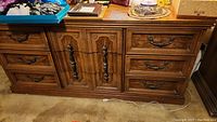 Long wooden dresser with nine drawers visible, center drawers create cabinet door illusion with metal handles and decorative carved wood details.
