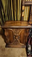 Photo of wooden night stand with ornate carved double-door cabinet and wood knobs against curtain background.