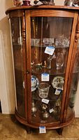 Front view of an oak wood display cabinet with a bowed glass front door and visible items inside on the glass shelves.