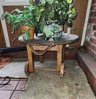 Image showing a rustic small round wooden table with two wooden wheels attached at the base and a thick pull rope tied between the legs. Several potted plants are placed on top of the table. The table sits on a carpeted floor near a brick wall.