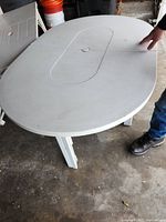 White oval patio table with folding top sections, supported by sturdy legs shown on concrete floor.