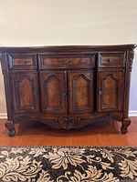 Front view of the wood buffet showing four cabinet doors and three drawers with decorative metal handles, ornate carvings on legs and base.