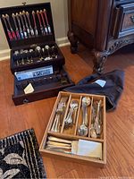 Photo showing two sets of flatware, one inside a wooden case and one in a tray, includes service pieces and napkin rings.