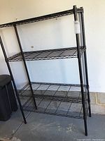 Front view of black metal wire shelving unit with four shelves and open wire grid design, standing against a gray floor and white wall.