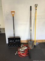 Snow shovel, dirt shovel, light bulb changing pole, extension cord, power strip, surge protector arranged against a wall.