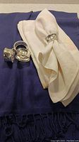 Four white linen napkins, some folded with one in a silver napkin ring, four silver napkin rings arranged on an indigo blue pashmina fabric with fringes.