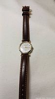 Top view of Alfred Sung ladies wrist watch with white dial, gold-tone case, and brown leather strap.