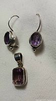 Pair of amethyst silver earrings and rectangular amethyst silver pendant necklace arranged on a white surface.