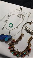 Photo showing assorted ladies jewelry on a white surface including fabric circle necklace, bead and star charm necklace, chain with bead accents, and green ring