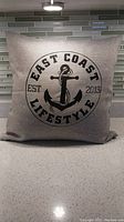 Front view of 12-inch square pillow with East Coast Lifestyle hand-screened anchor logo in black on a natural linen background, showing logo text and anchor graphic.