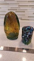 Two hand blown Canadian glass sculptures on a countertop with a ruler for scale. One is amber and green, about 9 inches tall, the other is cylindrical with a colorful swirling pattern, about 5 inches tall.