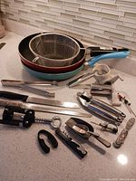 Photo showing two frying pans, frying basket, kitchen knives, cheese grater, garlic press, bottle opener, and various metal kitchen gadgets arranged on a kitchen counter.