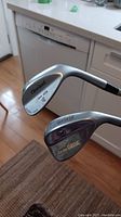 Photo showing two right-handed metal golf wedges held vertically. One features a Goliath brand with 55 degree loft, the other a Cleveland 588 RTX 64 degree wedge. Both appear to be in good shape with regripped handles.