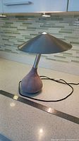 Full view of light grey suede bean bag base lamp with aluminum conical shade and power cord, placed on kitchen counter with ruler for scale.