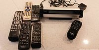 Roku SoundBridge streaming media device with power adapter and official remote plus six additional varied remote controls spread out on counter.