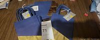 Photo showing multiple small gift bags, blue with gold bases, some with tags attached, placed on a wooden surface.