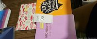 Photo showing a set of three items: a bright orange brain teaser book titled '3000 Puzzles, Riddles and Brain Teasers,' a pink Monday mini daily journal, and a cream journal with red lips pattern on the cover.