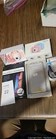 Wide view of the lot showing the pink digital camera in box, cell phone case with protective glass, Bluetooth earpiece, and packaging.