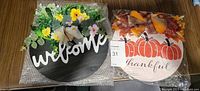Photo of two wooden home decor signs: one dark brown oval welcome sign with artificial yellow and white flowers and greenery, and one white round wooden sign with pumpkins, 'thankful' text, and fall-themed orange and red bow with leaves.