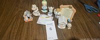 Seven figurines including a rocking donkey, unicorn, Precious Moments girl, angel bear, white house labeled 'Sugar,' seated bear, and a Christmas prayer plaque on a wooden floor.