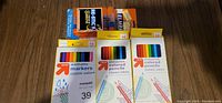 Photo of unopened new sets of colored pencils, washable markers, and Elmer's glue sticks arranged on a wooden surface.