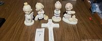 Five Precious Moments porcelain figurines displayed in a line with a white porcelain cross and auction card on a wooden surface.