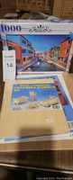 Photo showing the unopened 1000 piece puzzle box with a colorful image of a waterfront town, along with two dollhouse furniture kits in their original sealed packaging.