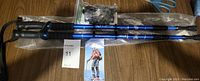 Pair of blue aluminum trekking poles lying on a table with black foam handles, wrist straps, and pointed tips with rubber covers, along with an instruction pamphlet and packaging visible.
