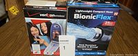 Two boxed items on a table: one is a Verizon Wireless flip phone box showing model LG VX6100, the other is a Bionic Flex lightweight compact hose box indicating 25 ft length, kink resistant and durable.