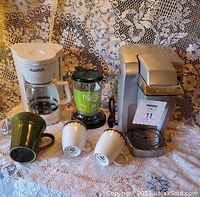 Coffee makers, tea steeper, and three mugs arranged on lace fabric background