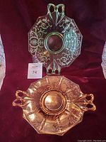 Both Fostoria two-handled plates, the green Vaseline glass above and amber-colored plate below, shown on red cloth background.