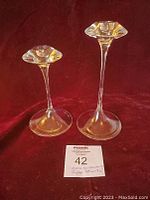 Two clear glass candle sticks on burgundy surface with a MaxSold number card.