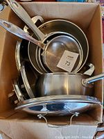 View of all five pots and four lids stacked in a cardboard box showing their size variations and condition.