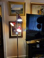 Full view of the standing lamp illuminated with the three red enamel light heads. Positioned next to a TV and framed paintings.