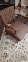 Image showing mid-century modern retro rocking chair with medium brown patterned upholstery and wooden armrests. Shows side view of chair on floral-patterned rug with other furniture in background.